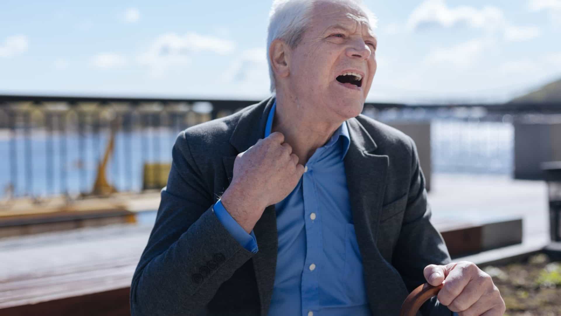 Homem idoso, de terno, puxando a gola para tentar respirar melhor