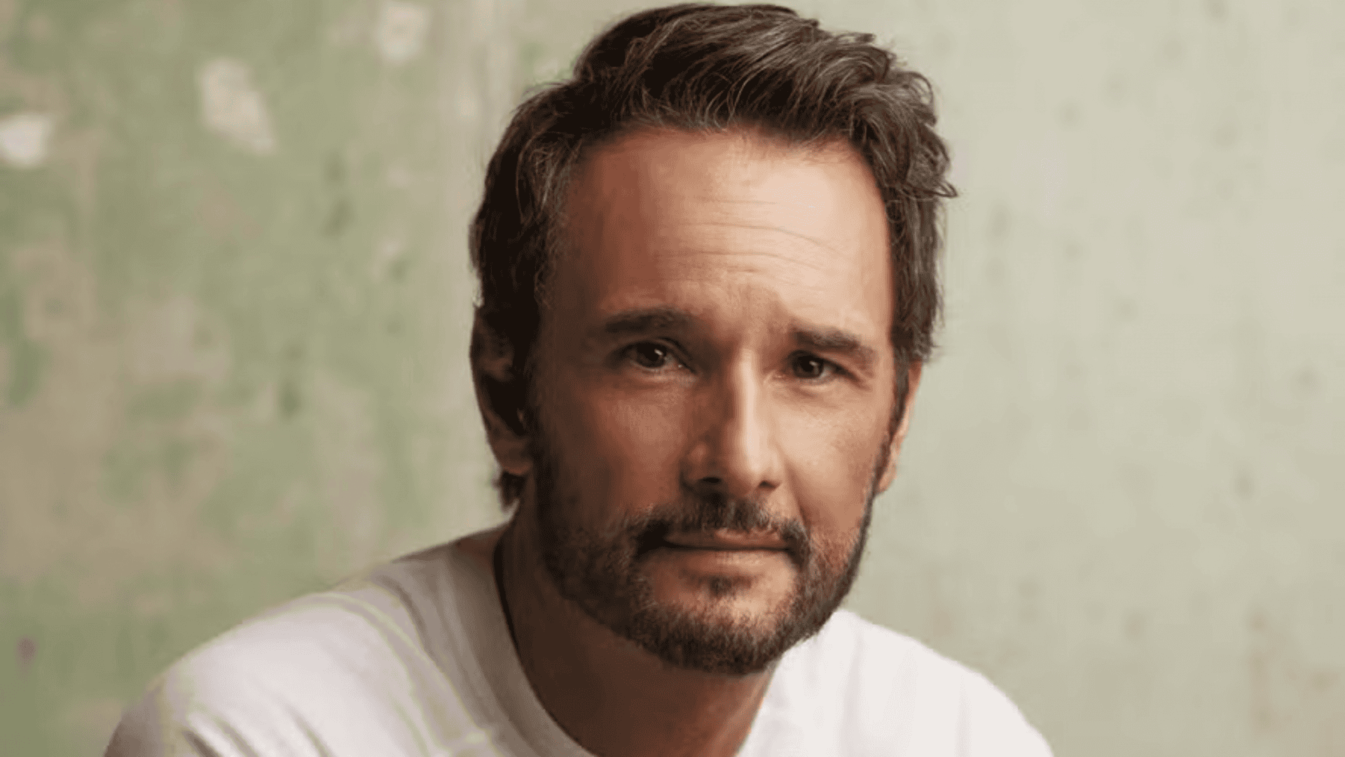 Rodrigo Santoro em um retrato próximo, vestindo uma camiseta branca. Ele tem barba curta e cabelos levemente ondulados, com expressão serena e olhar direto para a câmera. O fundo é uma parede texturizada em tom verde claro.