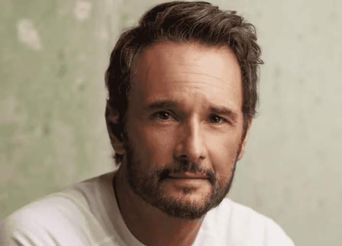 Rodrigo Santoro em um retrato próximo, vestindo uma camiseta branca. Ele tem barba curta e cabelos levemente ondulados, com expressão serena e olhar direto para a câmera. O fundo é uma parede texturizada em tom verde claro.