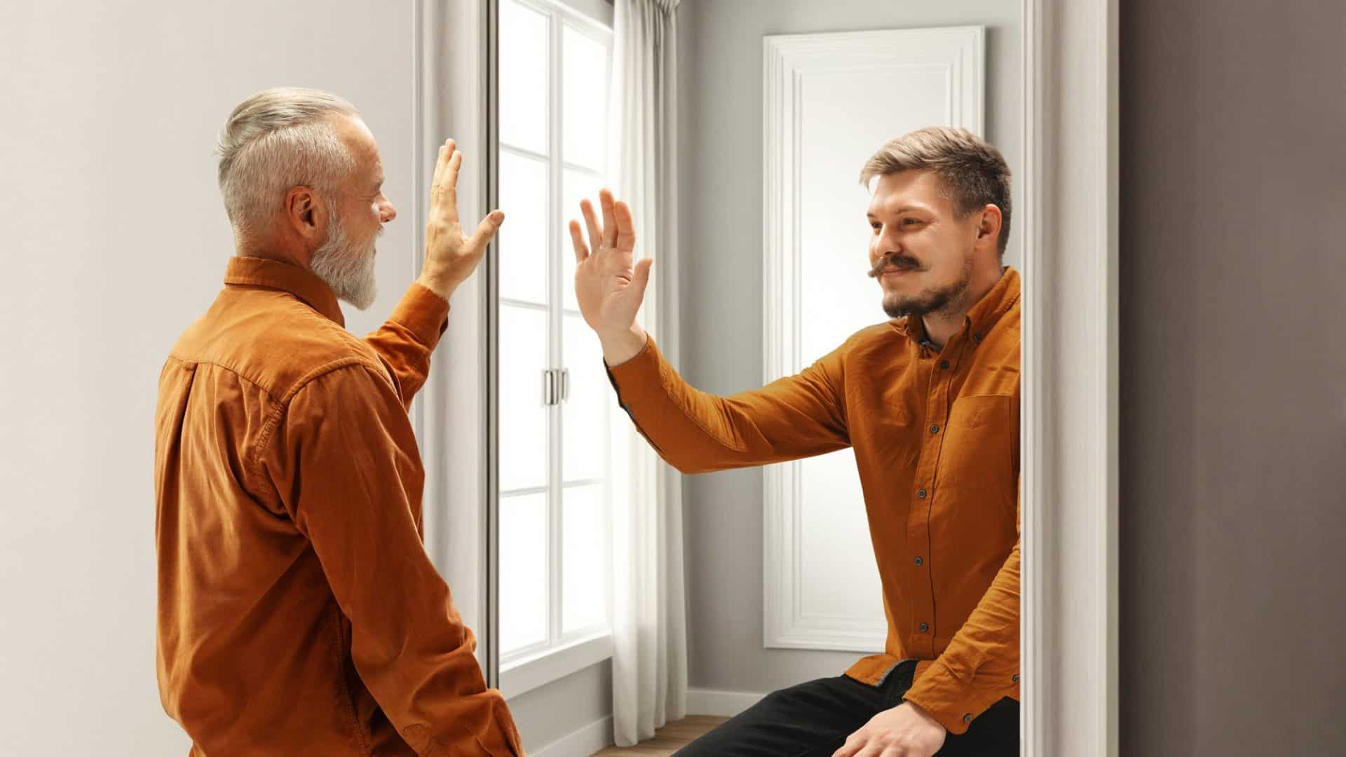 Dois homens se olhando no espelho e dando tchau um apra o outro, sendo que o da direita é uma versão mais velha do que se encontra à esquerda