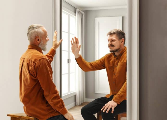 Dois homens se olhando no espelho e dando tchau um apra o outro, sendo que o da direita é uma versão mais velha do que se encontra à esquerda