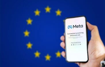 Ao fundo, desfocada, a bandeira da União Europeia; à direita, pessoa segurando um smartphone, no qual é exibida uma página da Meta