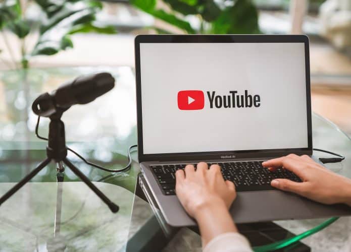 Do lado esquerdo, um microfone; do lado direito, uma pessoa mexendo em um notebook com o logo do YouTube em sua tela