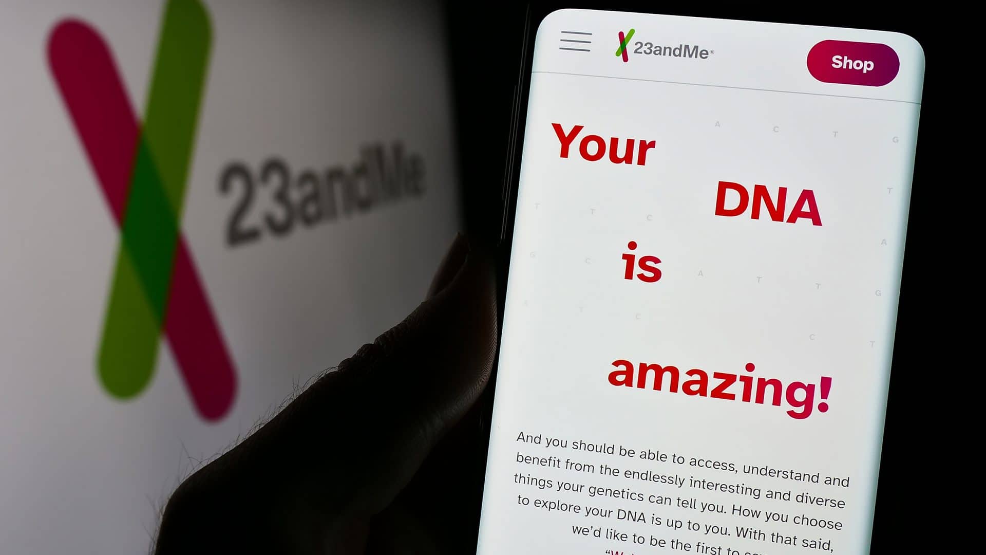 À esquerda, meio desfocado, logo da 23andMe de lado, em um monitor; à direita, uma pessoa segura um smartphone que exibe o site da empresa
