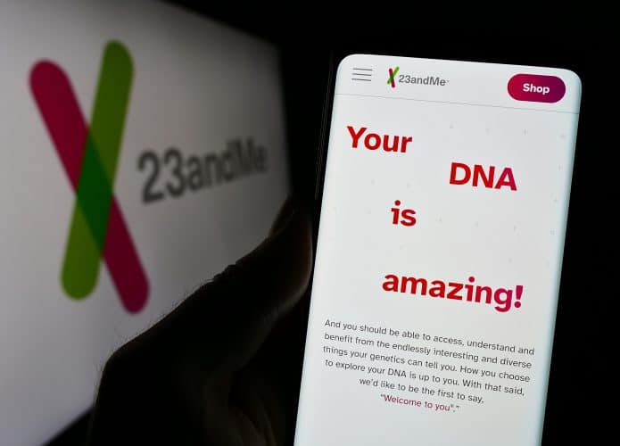 À esquerda, meio desfocado, logo da 23andMe de lado, em um monitor; à direita, uma pessoa segura um smartphone que exibe o site da empresa