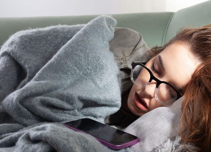 Jovem mulher exausta do vício do smartphone, dorme no sofá com seu telefone. Conceito de excesso de navegação nas redes sociais, uso excessivo da Internet e conexão on-line constante