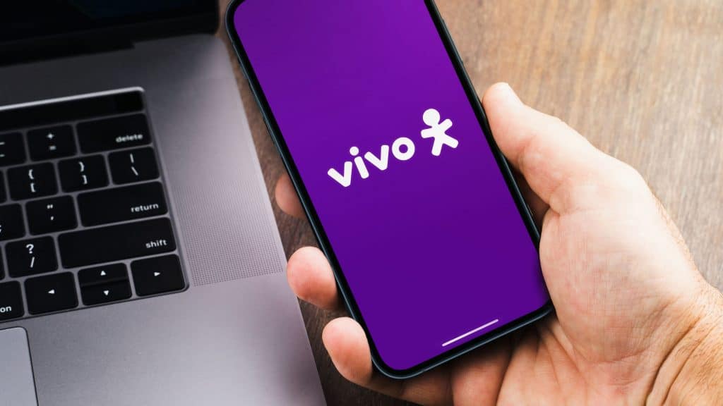 Pessoa segurando celular com logomarca da Vivo na Tela; ao lado, está um notebook