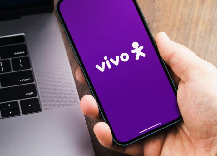 Pessoa segurando celular com logomarca da Vivo na Tela; ao lado, está um notebook
