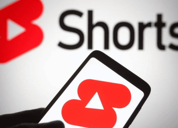 Pessoa segurando celular com logo do YouTube Shorts na tela, também exibido numa tela ao fundo