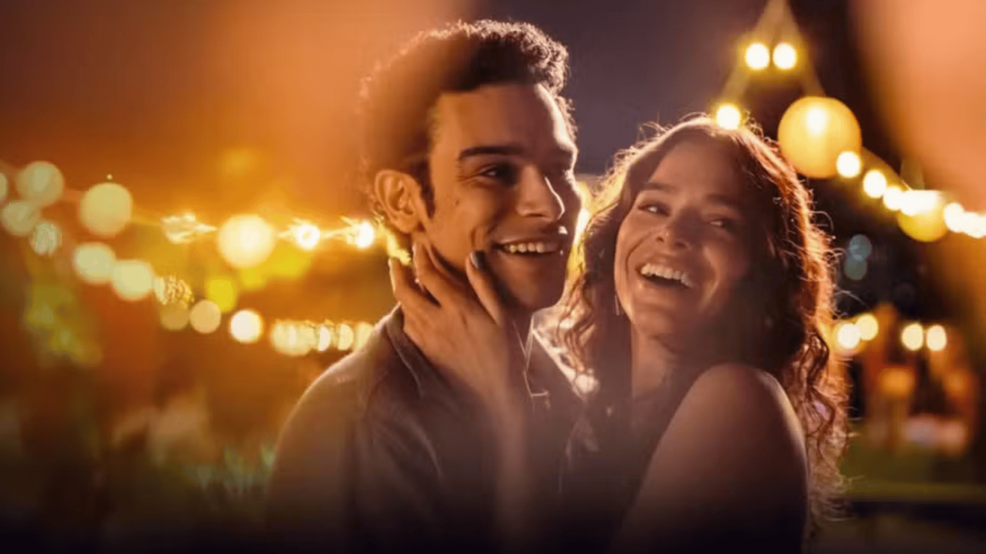 Cena noturna com um jovem casal sorridente em um ambiente iluminado por luzes decorativas e lanternas. A mulher segura o rosto do homem com carinho, e ambos parecem felizes