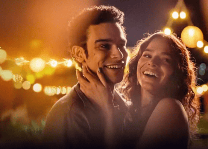 Cena noturna com um jovem casal sorridente em um ambiente iluminado por luzes decorativas e lanternas. A mulher segura o rosto do homem com carinho, e ambos parecem felizes