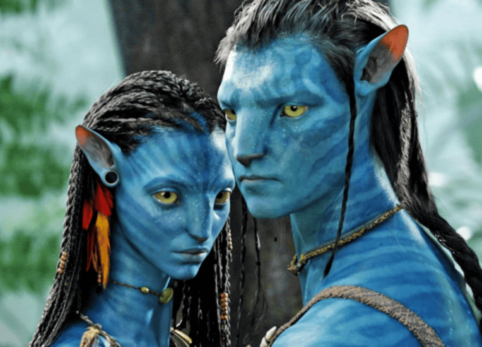 Cena do filme Avatar com dois personagens da espécie Na’vi, de pele azul e olhos amarelos, em um ambiente de floresta exuberante. Eles estão próximos, com expressões sérias e detalhes tribais no rosto e vestimentas.