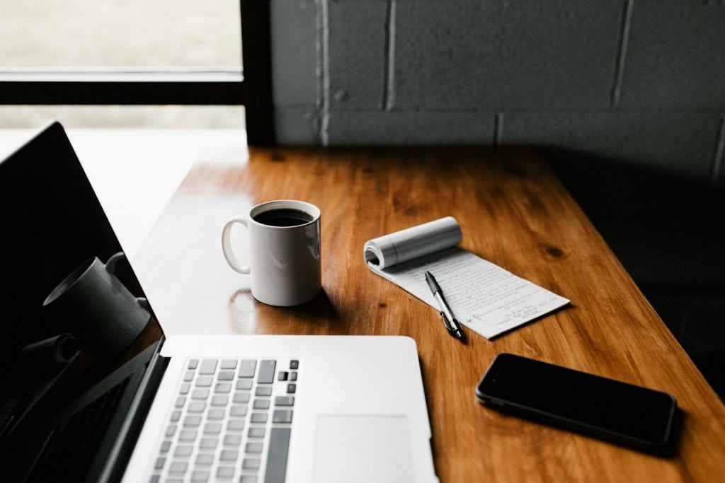 computador aberto em cima de uma mesa com xícara de café e bloco de anotações