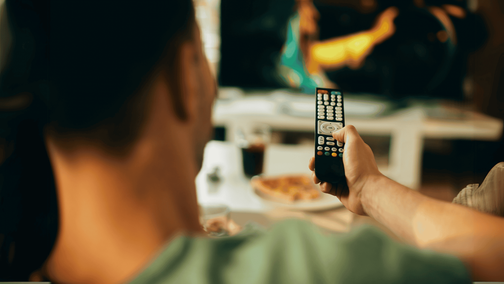 Pessoa segura um controle remoto enquanto assiste à TV. A cena é vista de trás, com a televisão ao fundo exibindo uma imagem desfocada. Na mesa à frente, há um prato de pizza e um copo de refrigerante