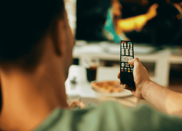 Pessoa segura um controle remoto enquanto assiste à TV. A cena é vista de trás, com a televisão ao fundo exibindo uma imagem desfocada. Na mesa à frente, há um prato de pizza e um copo de refrigerante