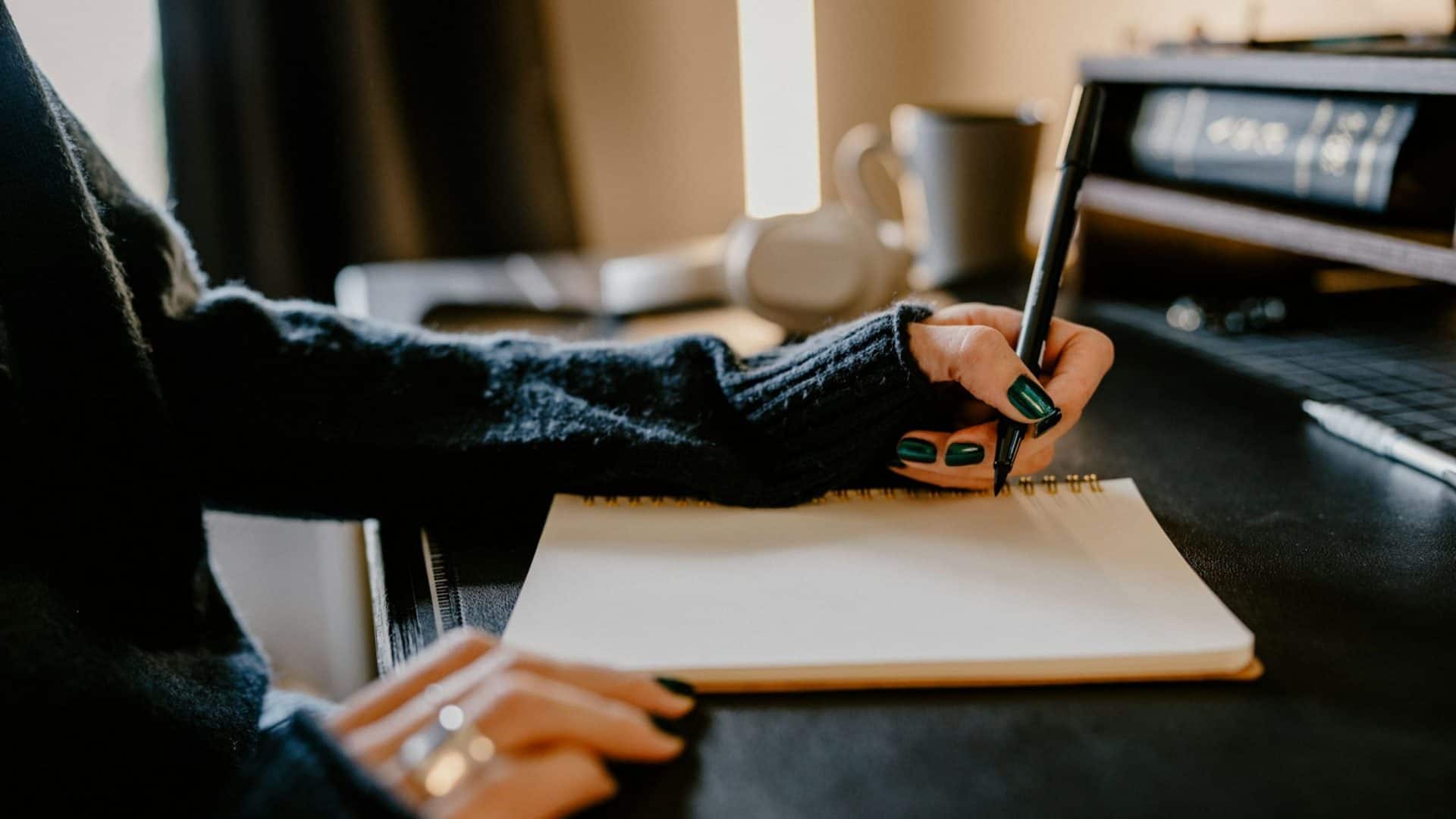 mulher com blusa de frio escura e unhas pretas escrevendo em um caderno