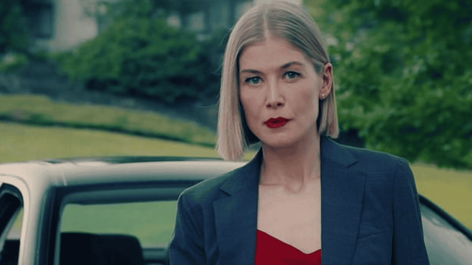 Rosamund Pike com um olhar determinado, vestindo um blazer azul-escuro sobre uma blusa vermelha, com batom da mesma cor. Ao fundo, um carro e uma paisagem verde desfocada