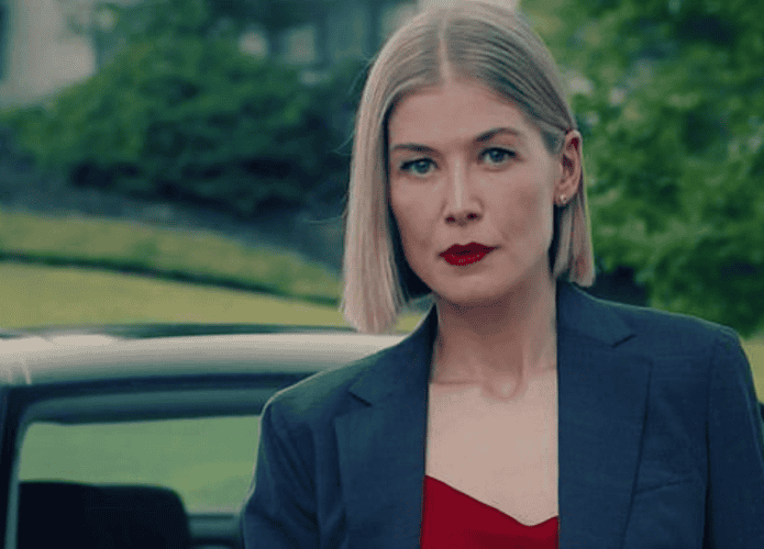 Rosamund Pike com um olhar determinado, vestindo um blazer azul-escuro sobre uma blusa vermelha, com batom da mesma cor. Ao fundo, um carro e uma paisagem verde desfocada