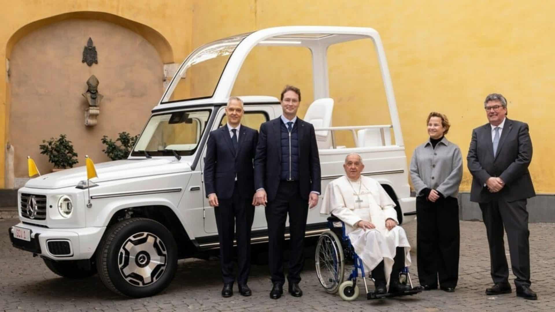 Papa Francisco em cadeira de rodas ao lado de executivos da Mercedes com papamóvel elétrico atrás deles