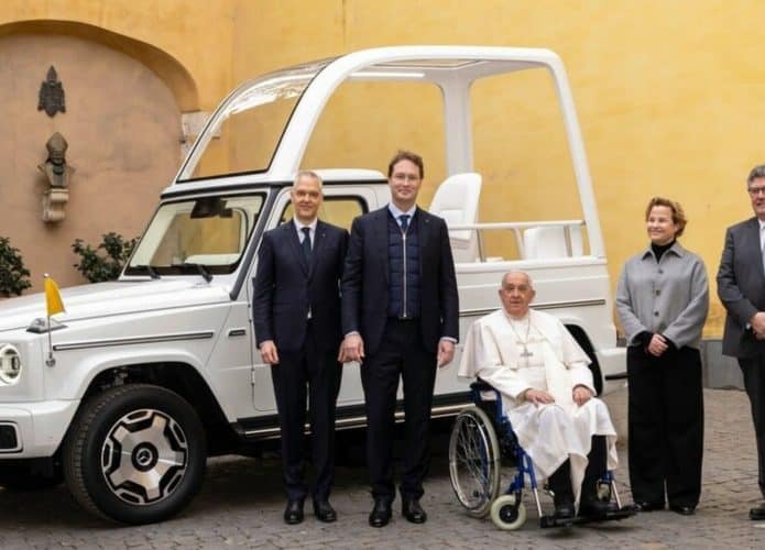 Papa Francisco em cadeira de rodas ao lado de executivos da Mercedes com papamóvel elétrico atrás deles