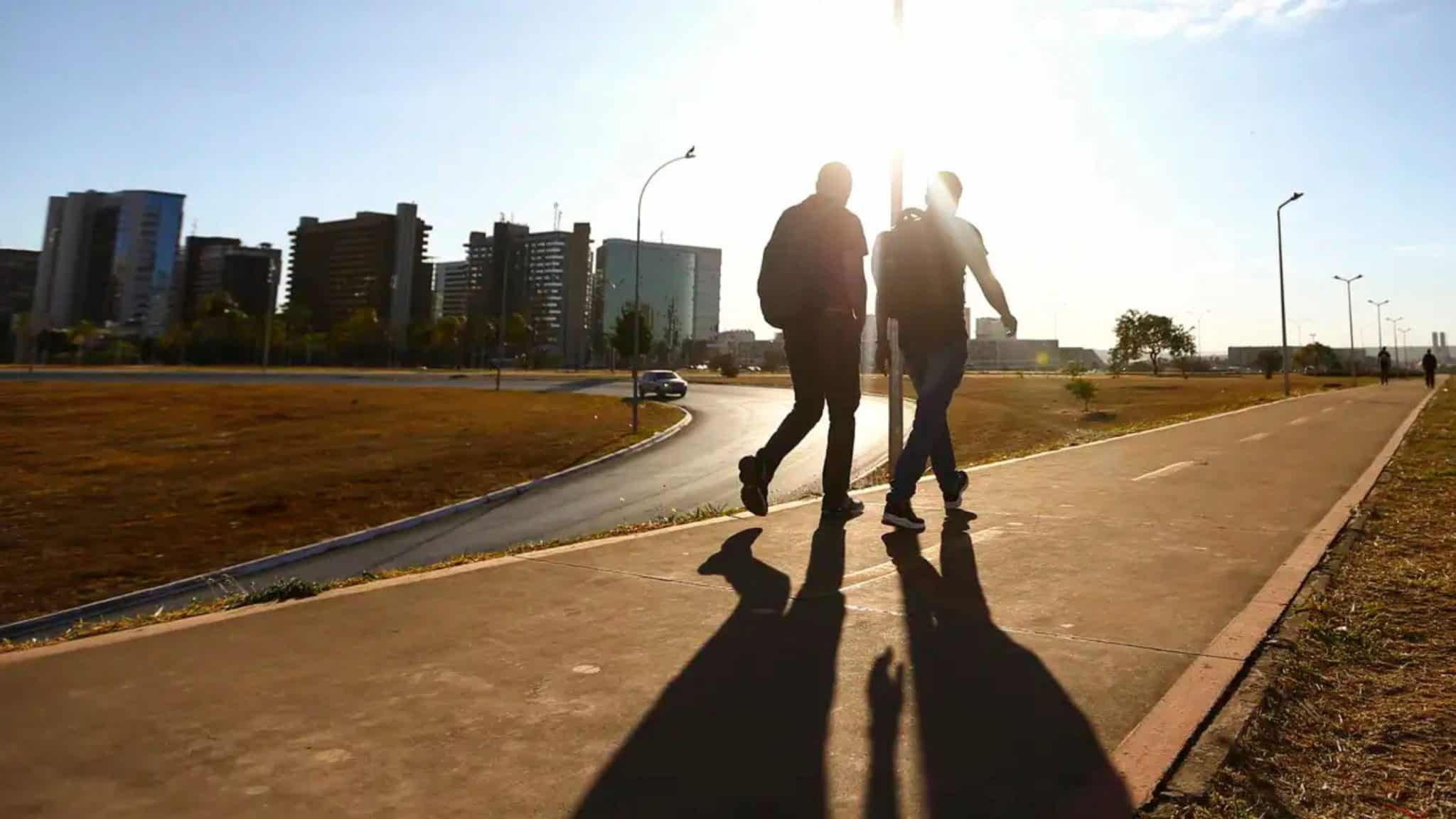 Duas pessoas caminhando sob sol de outono em calçada de avenida