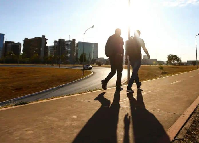 Duas pessoas caminhando sob sol de outono em calçada de avenida