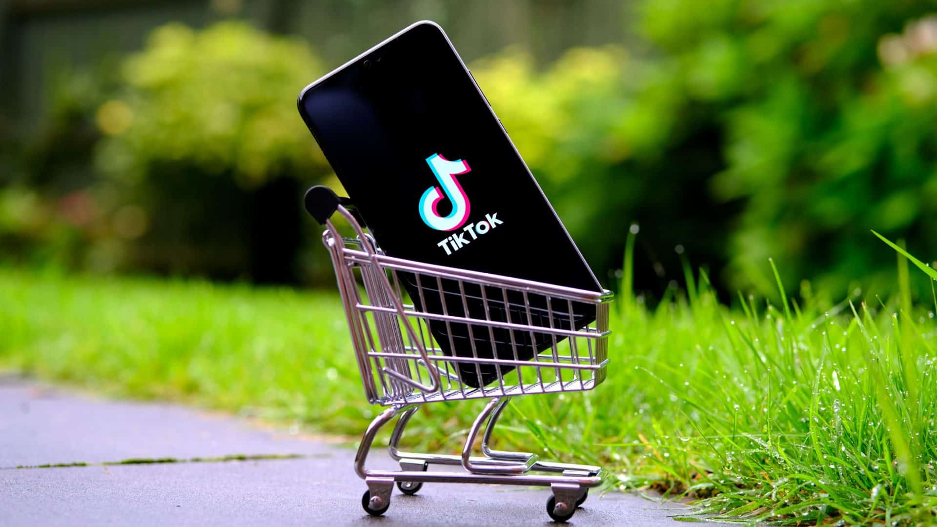 Celular com logomarca do TikTok na tela dentro de miniatura de carrinho de mercado numa calçada com gramado no fundo