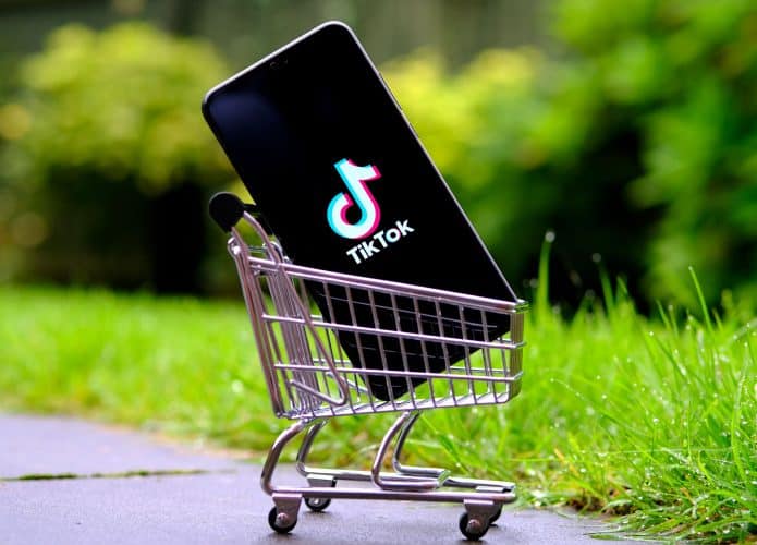 Celular com logomarca do TikTok na tela dentro de miniatura de carrinho de mercado numa calçada com gramado no fundo