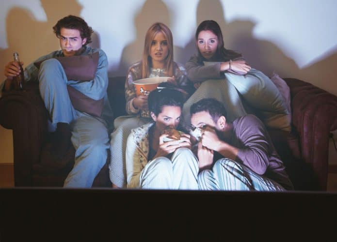 imagem mostra amigos adultos reunidos na sala de casa, assistindo a um filme de terror ação