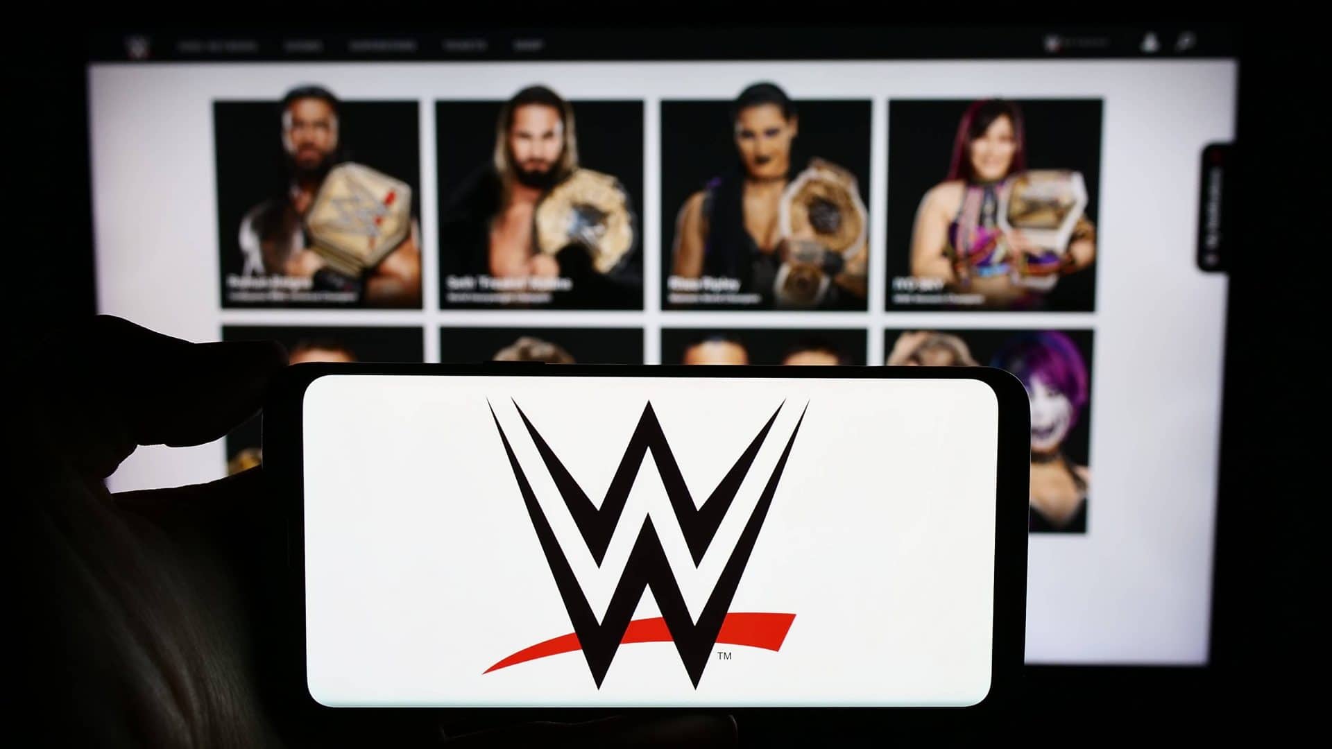 imagem mostra um celular com a logo das lutas WWE em frente de um monitor com vários lutadores no fundo