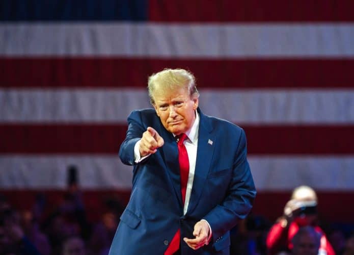 Donald Trump apontando para a frente; atrás, parte da bandeira dos EUA desfocada