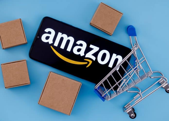 Logo da Amazon em um smartphone, que está em um mini carrinho de compras e duas mini caixas