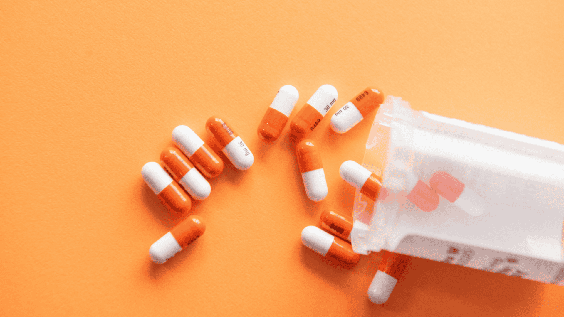 imagem mostra um frasco plástico sobre uma tela laranja, despejando várias unidades de pílulas medicamentosas