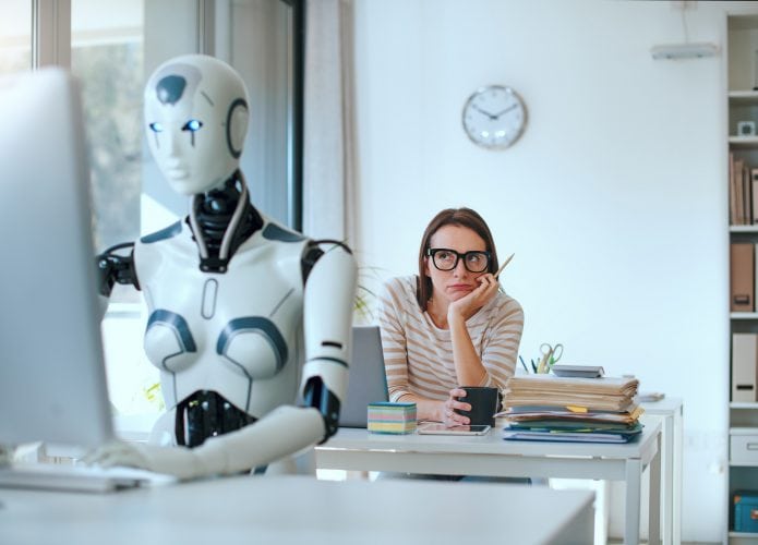 Robô sentado em frente a um computador, com uma mulher emburrada atrás olhando para ele