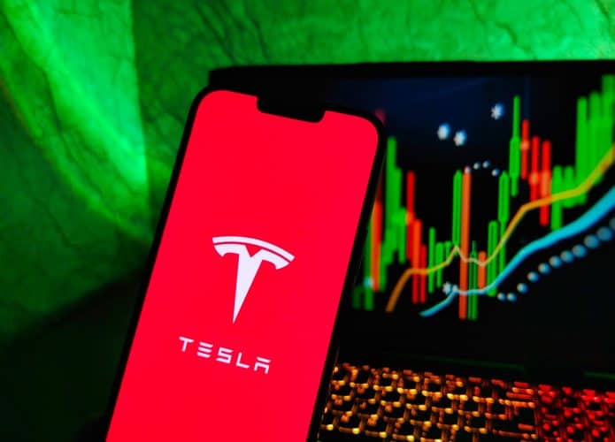 À frente, logo da Tesla em um smartphone; ao fundo, um notebook com gráficos