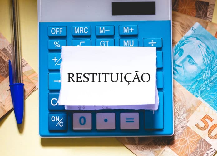 A palavra restituição em português do Brasil escrita em um pedaço de papel sobre uma calculadora. Notas de Real brasileiro na composição. Economia brasileira e imposto de renda.