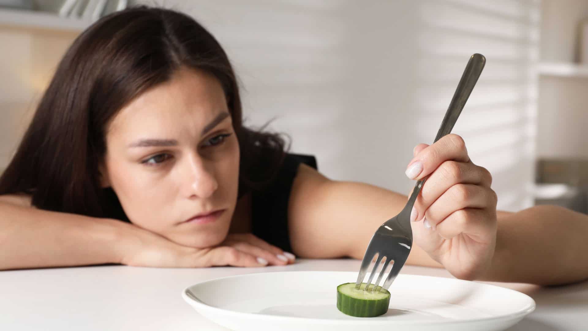 Transtorno alimentar. Mulher triste segurando garfo com pepino à mesa em ambiente fechado