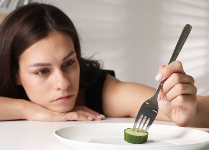 Transtorno alimentar. Mulher triste segurando garfo com pepino à mesa em ambiente fechado