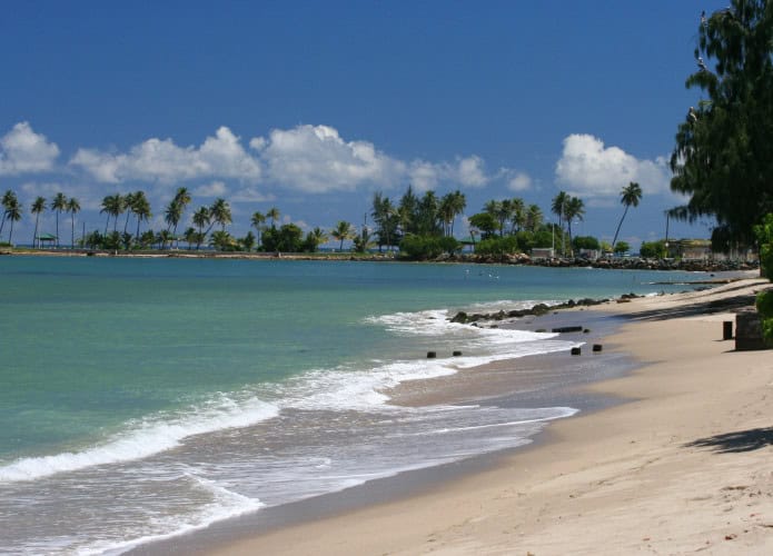 Praia de Luquillo em Porto Rico com águas azul-turquesa, faixa de areia clara e palmeiras ao fundo sob um céu azul com nuvens.