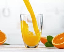 Suco de laranja sendo despejado em copo grande de vidro rodeado de laranjas cortadas