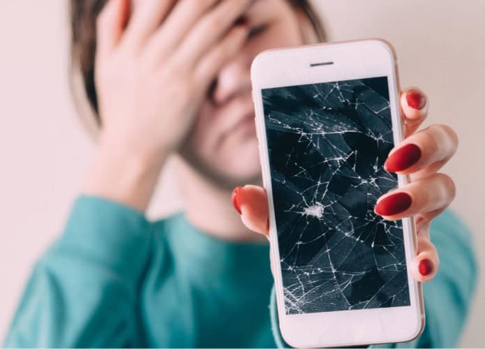 imagem mostra uma adolescente chateada enquanto segura na frente da câmera um iphone com a tela quebrada