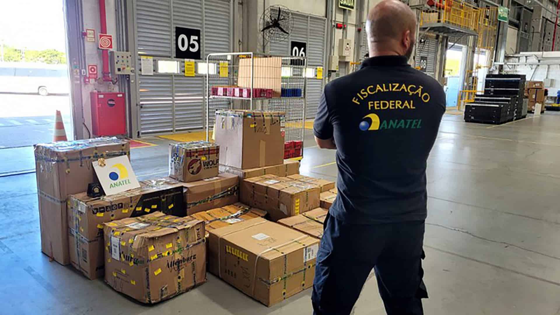 Várias caixas de papelão lacradas e empilhadas, com um agente da Anatel de costas