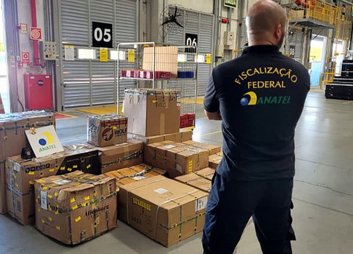 Várias caixas de papelão lacradas e empilhadas, com um agente da Anatel de costas