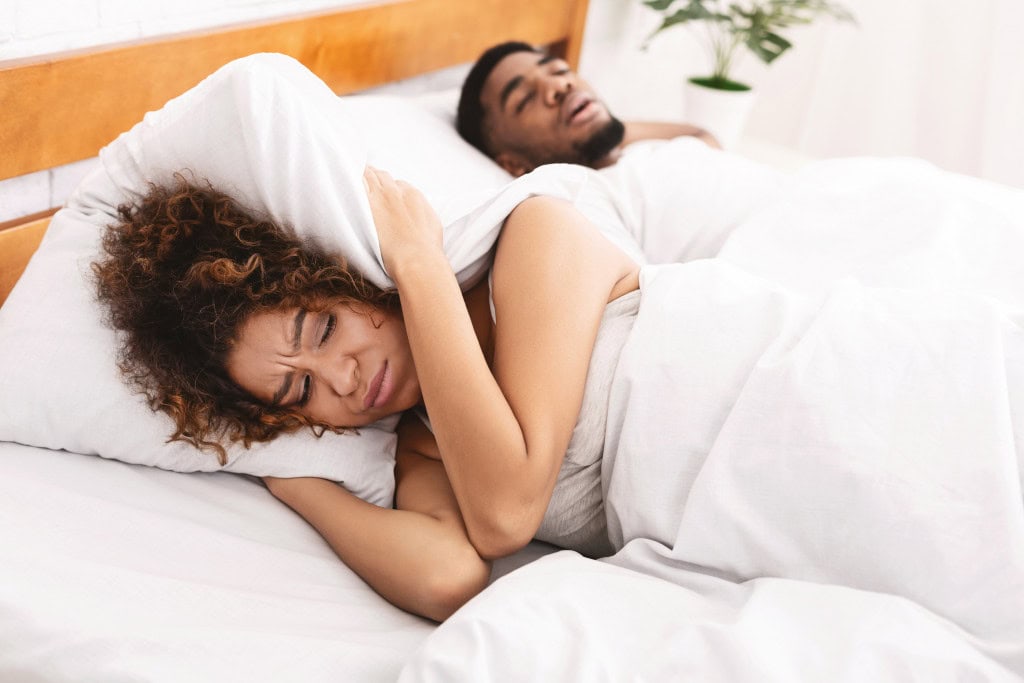 Mulher tampando os ouvidos com o travesseiro ao lado de um homem que está roncando