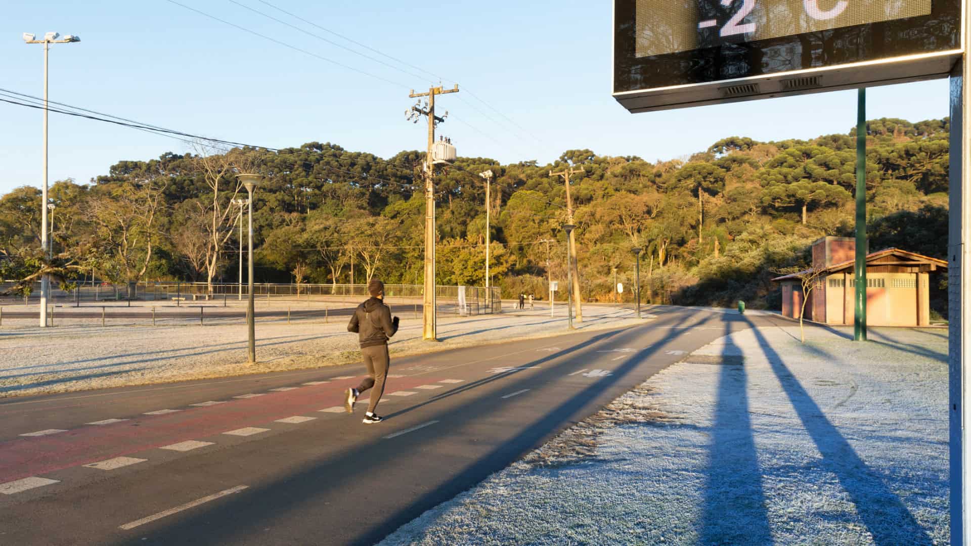 Estrada ensolarada com neve na lateral direita da foto; acima, um termômetro de rua marcando −2 °C