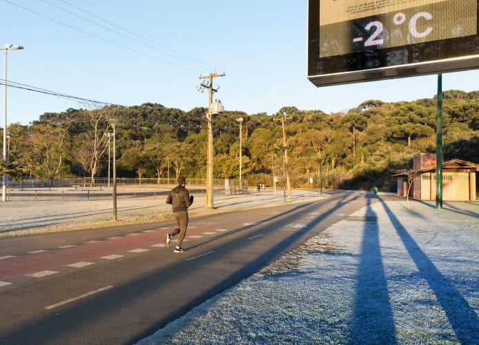 Estrada ensolarada com neve na lateral direita da foto; acima, um termômetro de rua marcando −2 °C