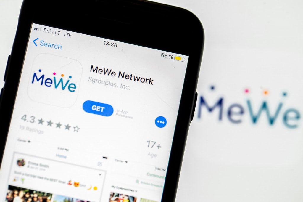 Logo do MeWe ao fundo e desfocado; à frente, página de download do MeWe na App Store exibida em um iPhone