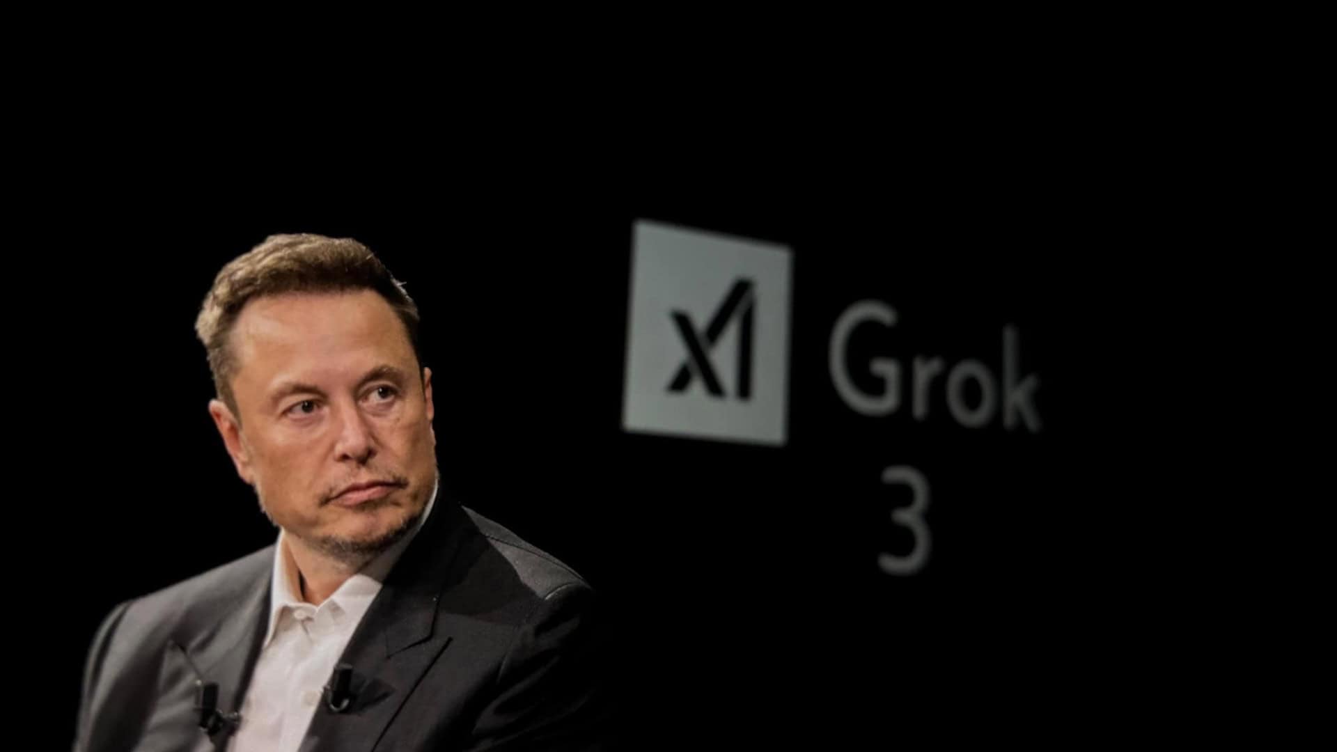 Elon Musk ao lado de uma montagem com os logos de xAI e Grok