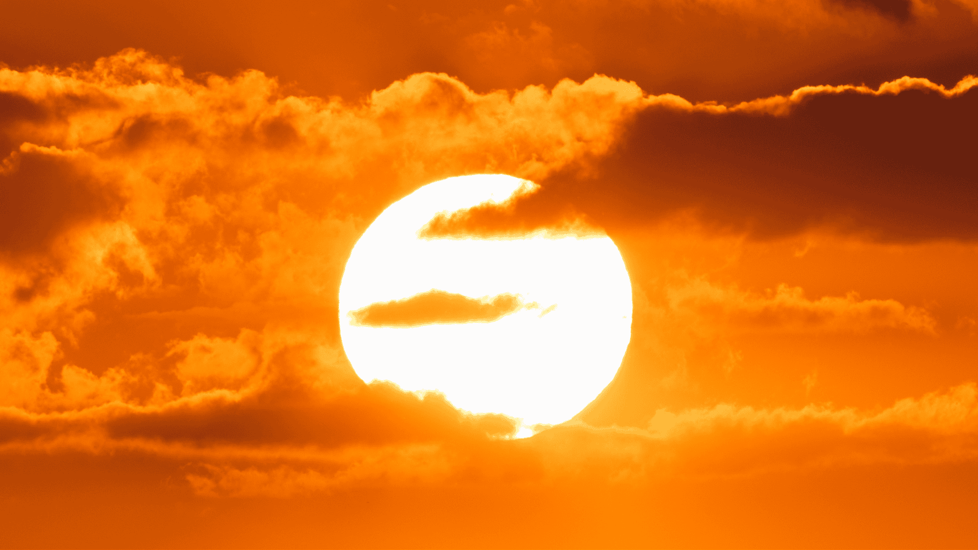 Imagem do Sol brilhando intensamente entre nuvens alaranjadas durante o pôr do sol, com céu em tons quentes