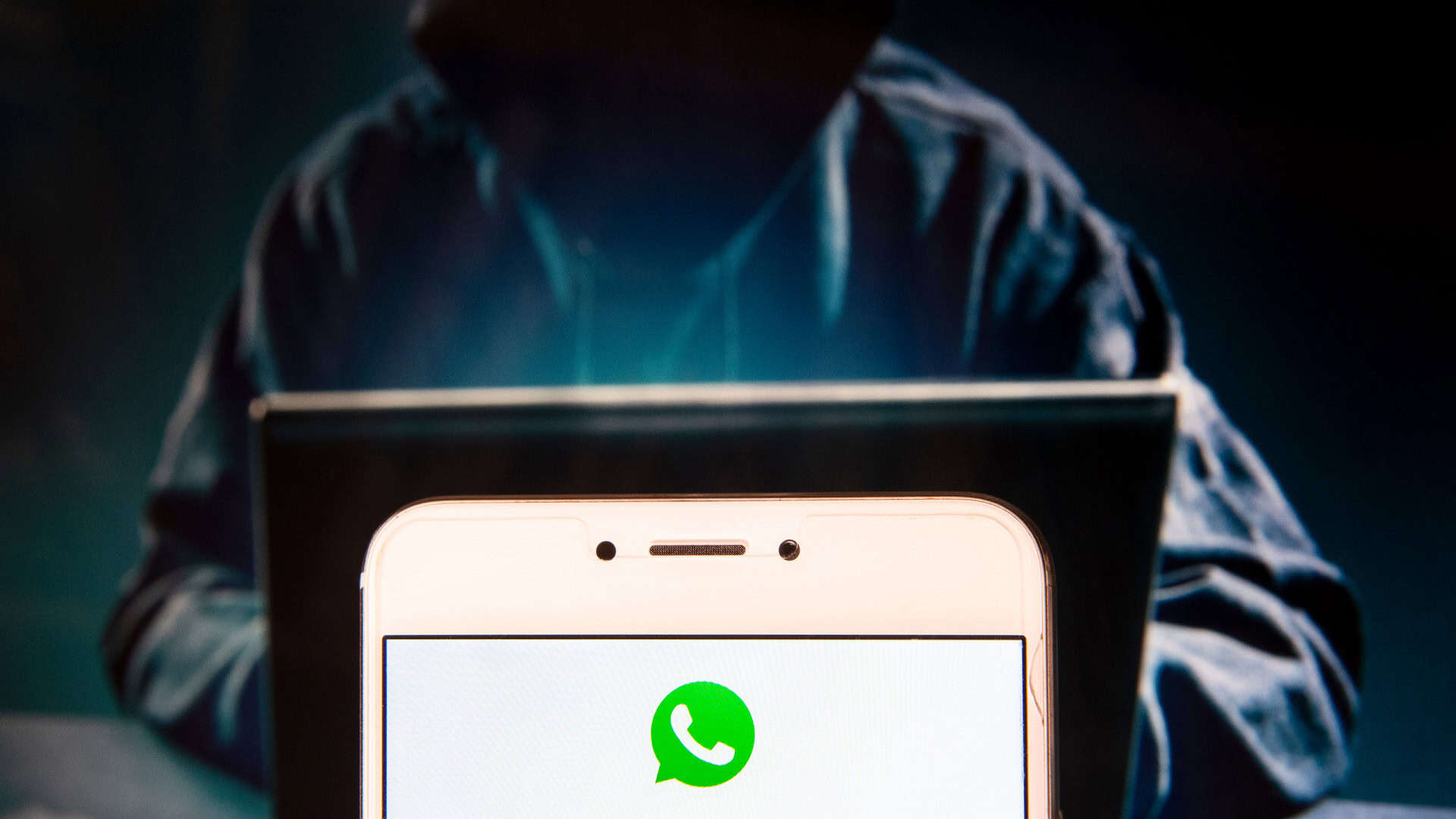 Pessoa com capuz à frente de um notebook; em primeiro plano, logo do WhatsApp em um smartphone